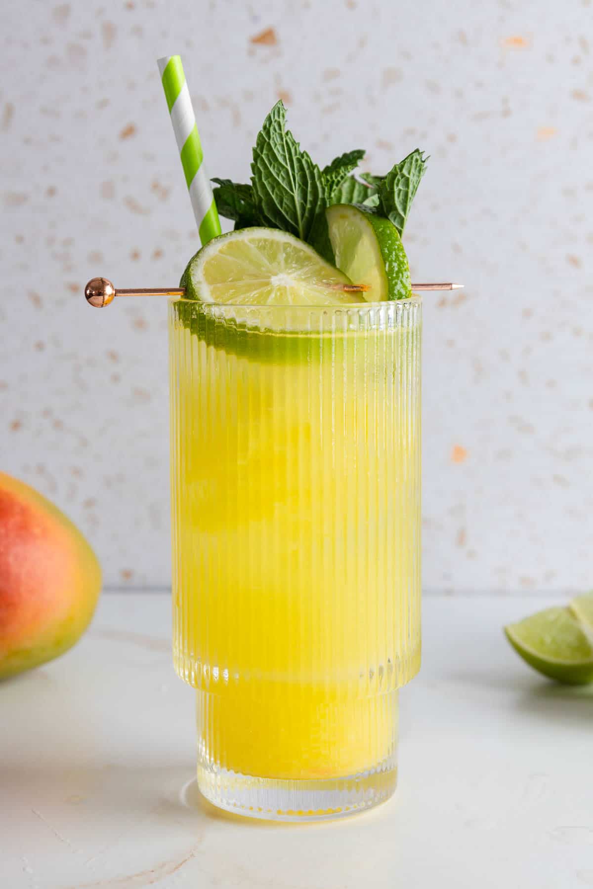 A yellow mango tropical drink with ice, mint, lime slices, and a striped straw in a glass on a white surface&mdash;a refreshing mocktail recipe perfect for summer.