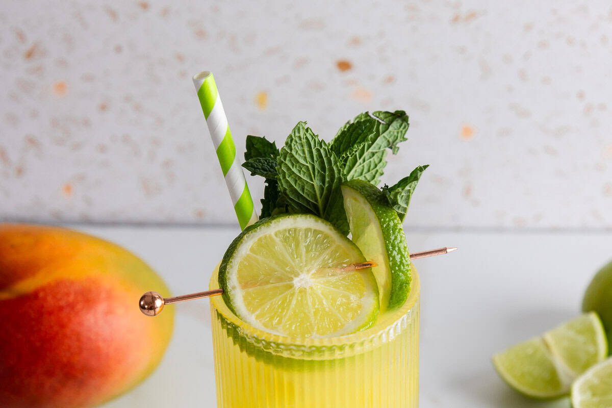 Refreshing yellow electrolyte mocktail recipe with lime slice, mint leaves, and striped straw, garnished with mango and lime in the background.