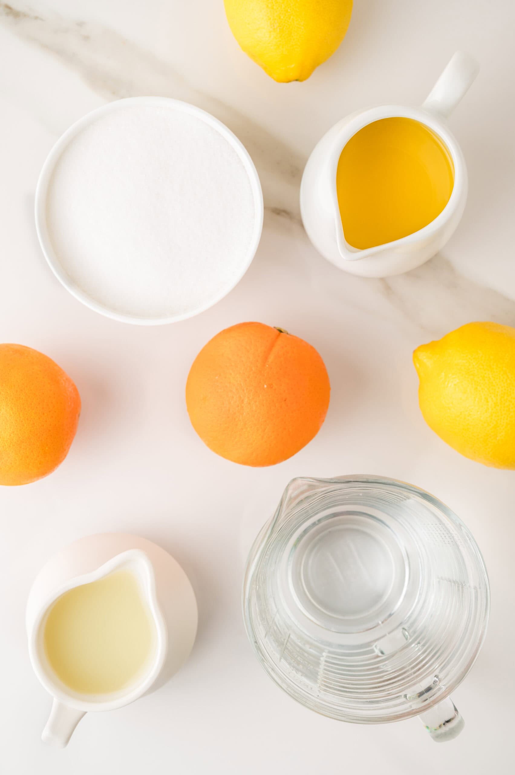 Overhead view of oranges, lemons, sugar, water, and small pitchers of juice and milk on a white surface&mdash;perfect ingredients for a refreshing homemade orangeade recipe.