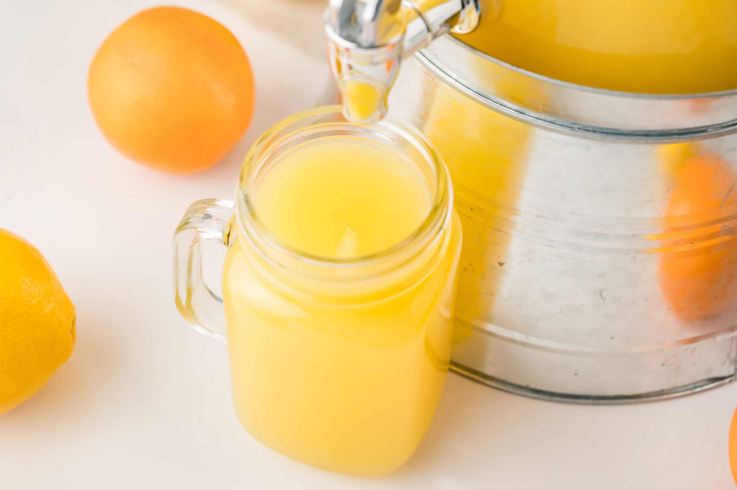 A glass jar is filled with homemade orangeade from a metal dispenser, surrounded by oranges and a lemon.