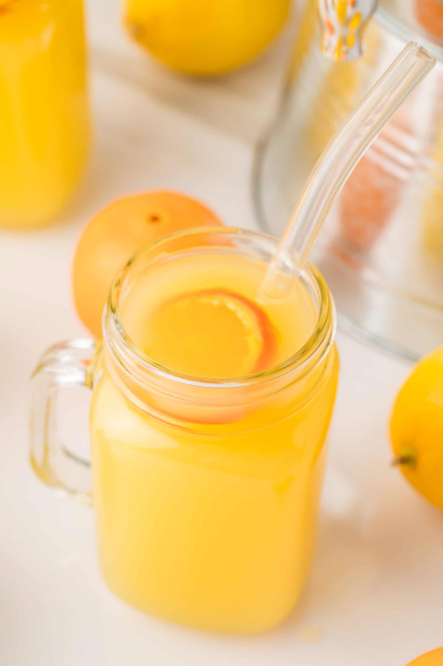 A glass mug of homemade orange drink with a straw and an orange slice, surrounded by fresh oranges and lemons.