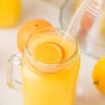 A mason jar of homemade orangeade with a lemon slice and glass straw, surrounded by fresh lemons.
