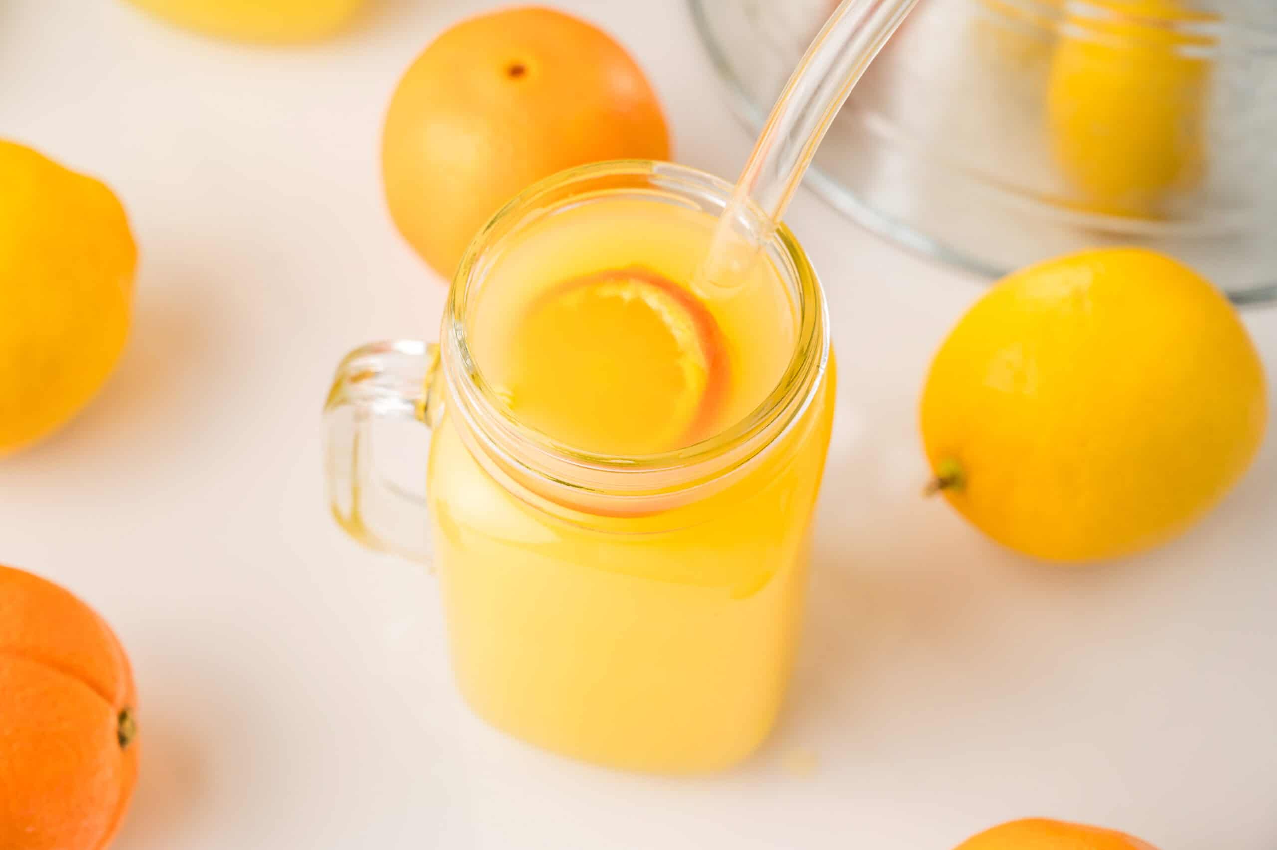 A glass mug of Homemade Orange Drink with a straw, surrounded by whole oranges and lemons on a white surface.