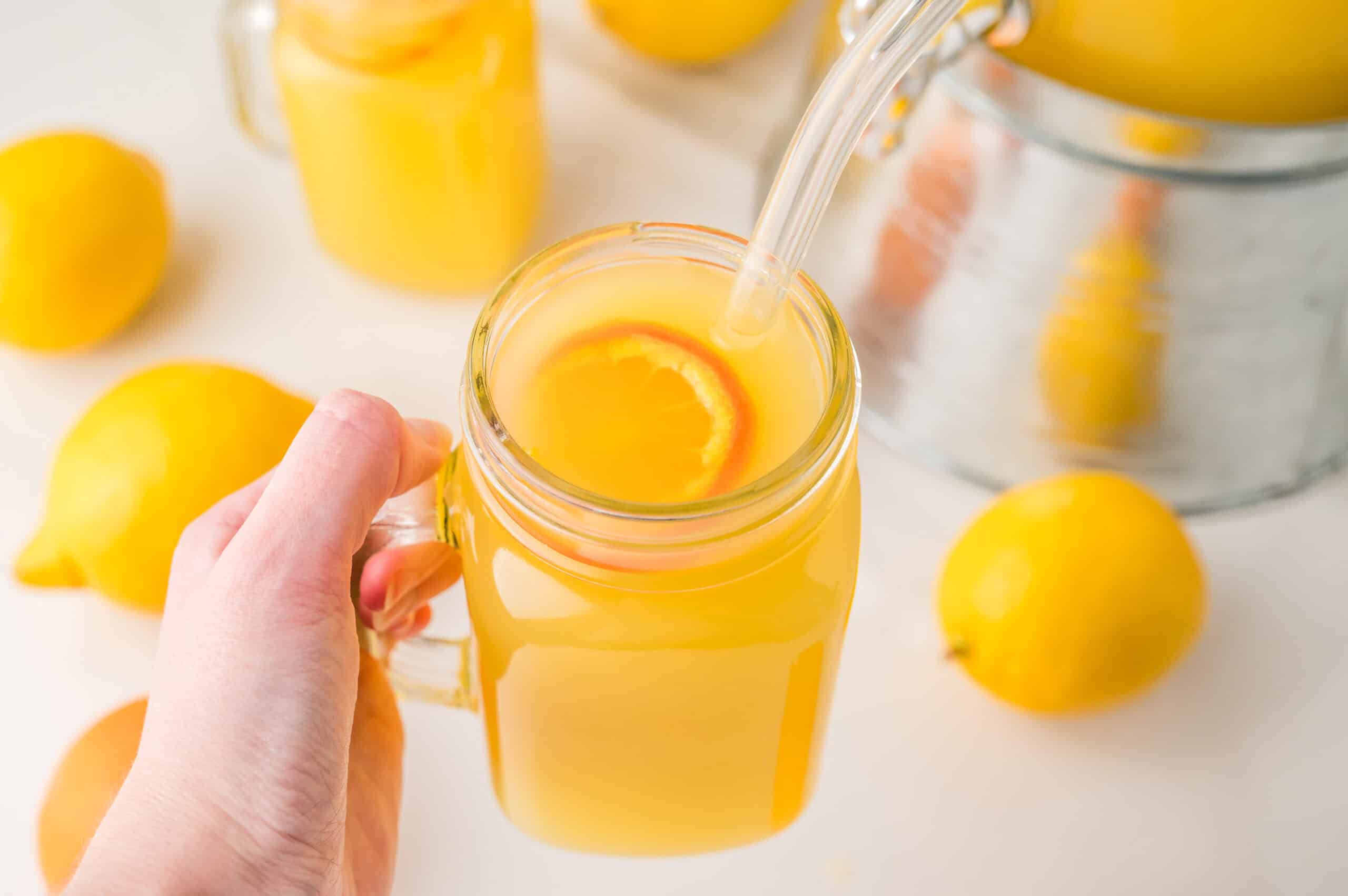 A hand holds a mason jar of lemonade with a straw and lemon slice, surrounded by fresh lemons&mdash;perfect inspiration for trying your own Homemade Orangeade or favorite orangeade recipe.