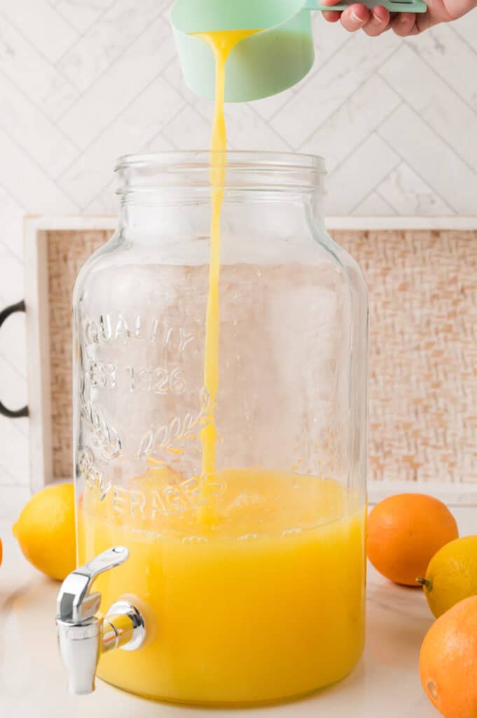 A hand pours homemade orange drink from a cup into a large glass dispenser surrounded by whole oranges.