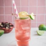 A pink pomegranate drink with ice, lime garnish, and a striped straw in a glass, set against a pink tiled background—perfect as an electrolyte-packed mocktail recipe.