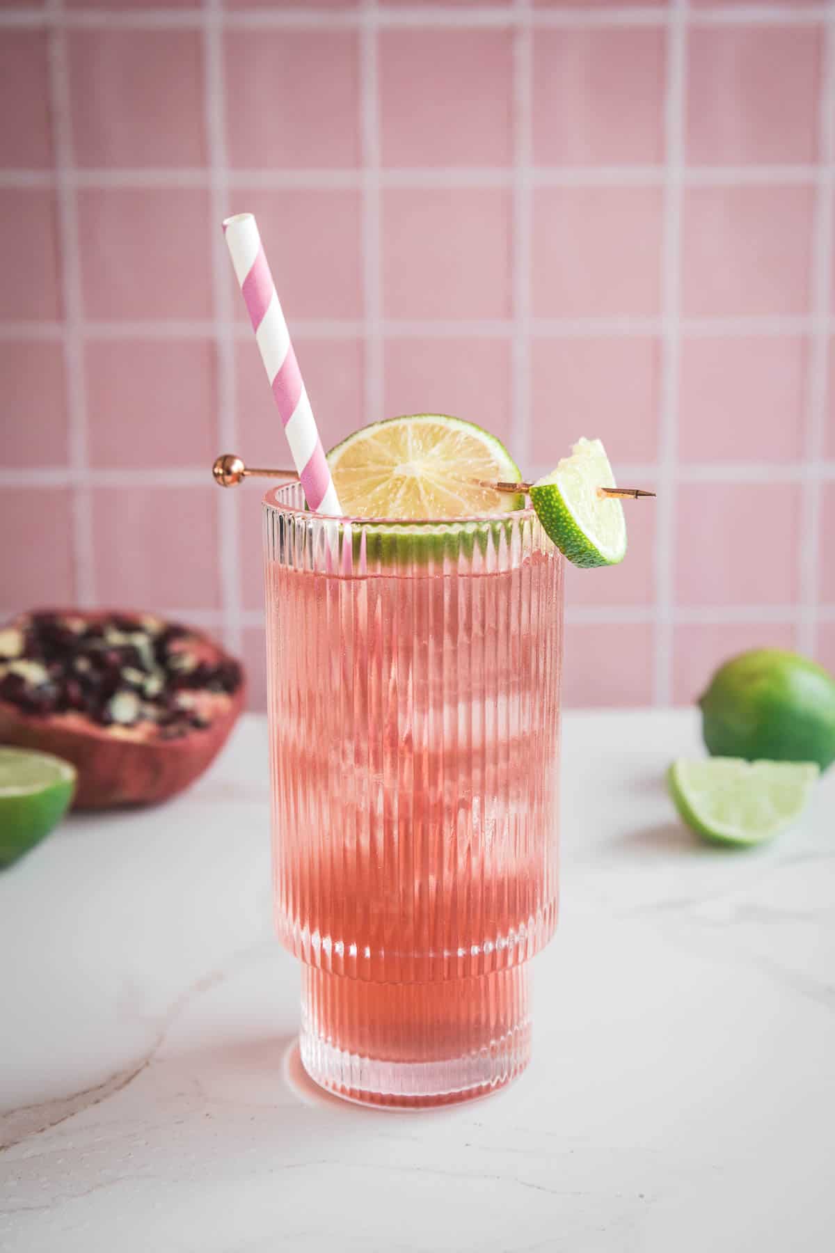 A pink pomegranate drink with ice, lime garnish, and a striped straw in a glass, set against a pink tiled background&mdash;perfect as an electrolyte-packed mocktail recipe.