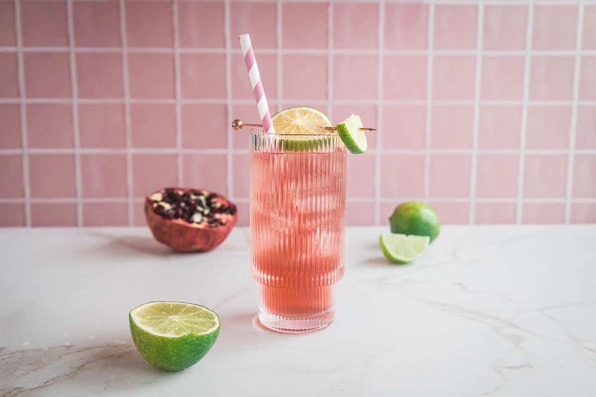 A pink pomegranate mocktail with ice, lime and lemon slices, and a striped straw sits on a marble counter with fruit nearby&mdash;an energizing electrolyte boost in every sip.