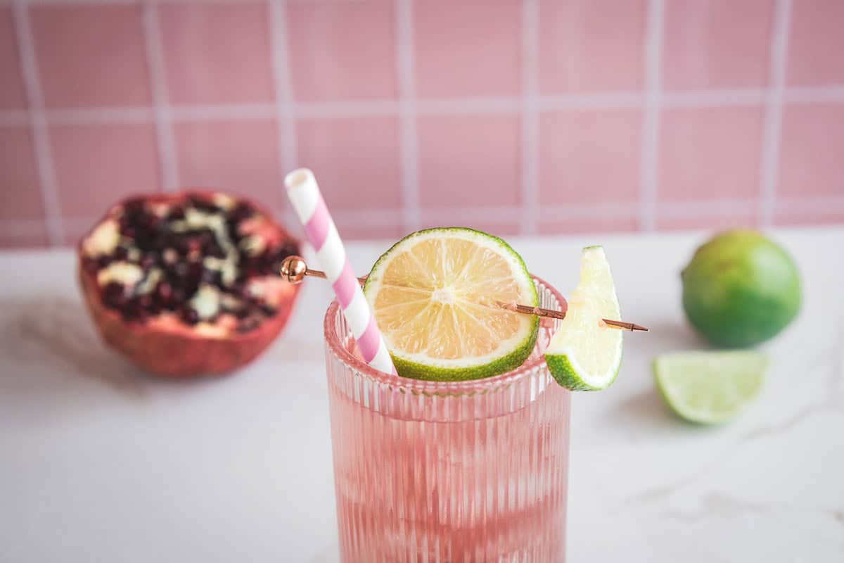 Pink electrolyte drink in a glass with a striped straw and lime slices, with pomegranate and limes in the background&mdash;an irresistible mocktail recipe for any occasion.