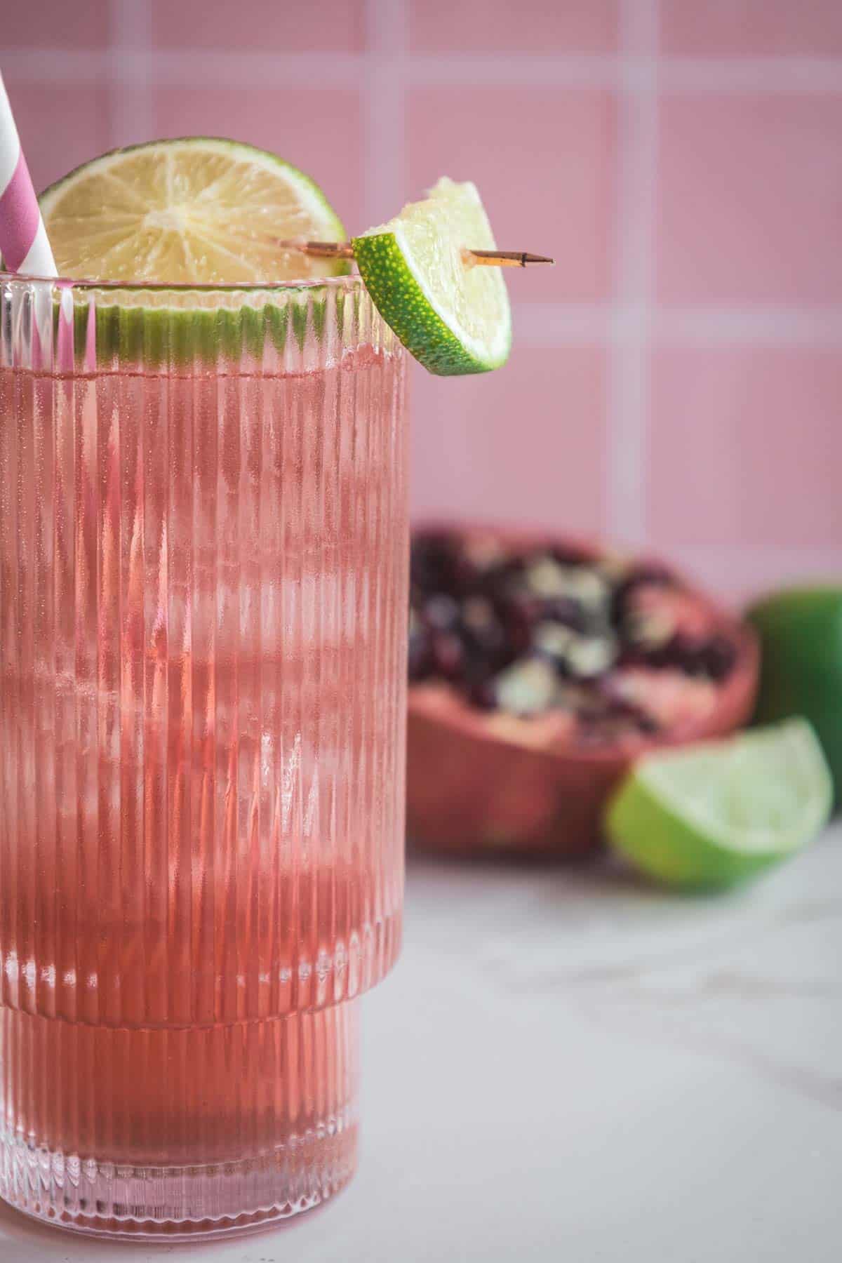 A pink electrolyte mocktail in a ribbed glass with ice, a lime garnish, and a striped straw; pomegranate and lime blurred behind&mdash;an inviting mocktail recipe for refreshing hydration.