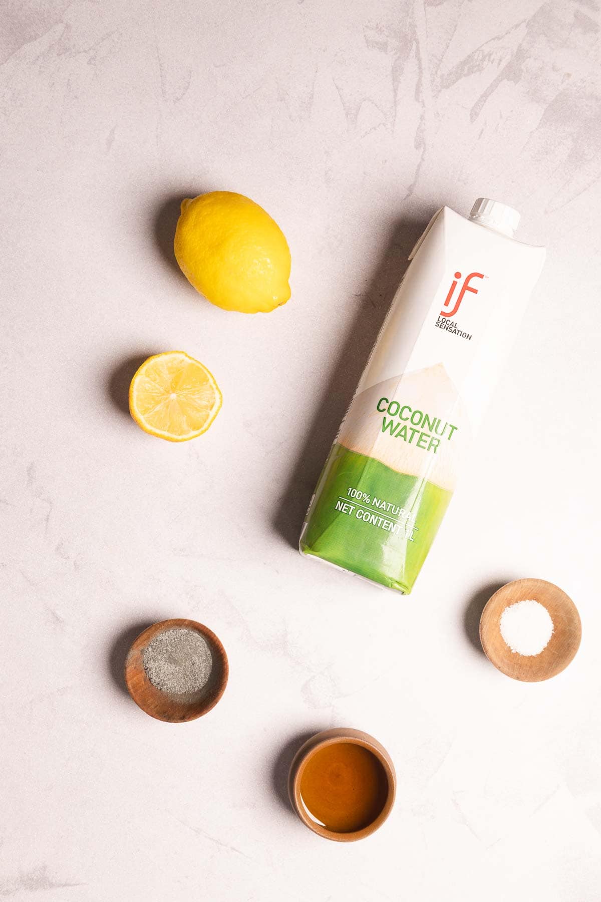 Whole lemon, half lemon, coconut water carton, bowls of pepper and salt, plus a small bowl of honey on a white surface&mdash;perfect ingredients for a refreshing Electrolyte Mocktail recipe.