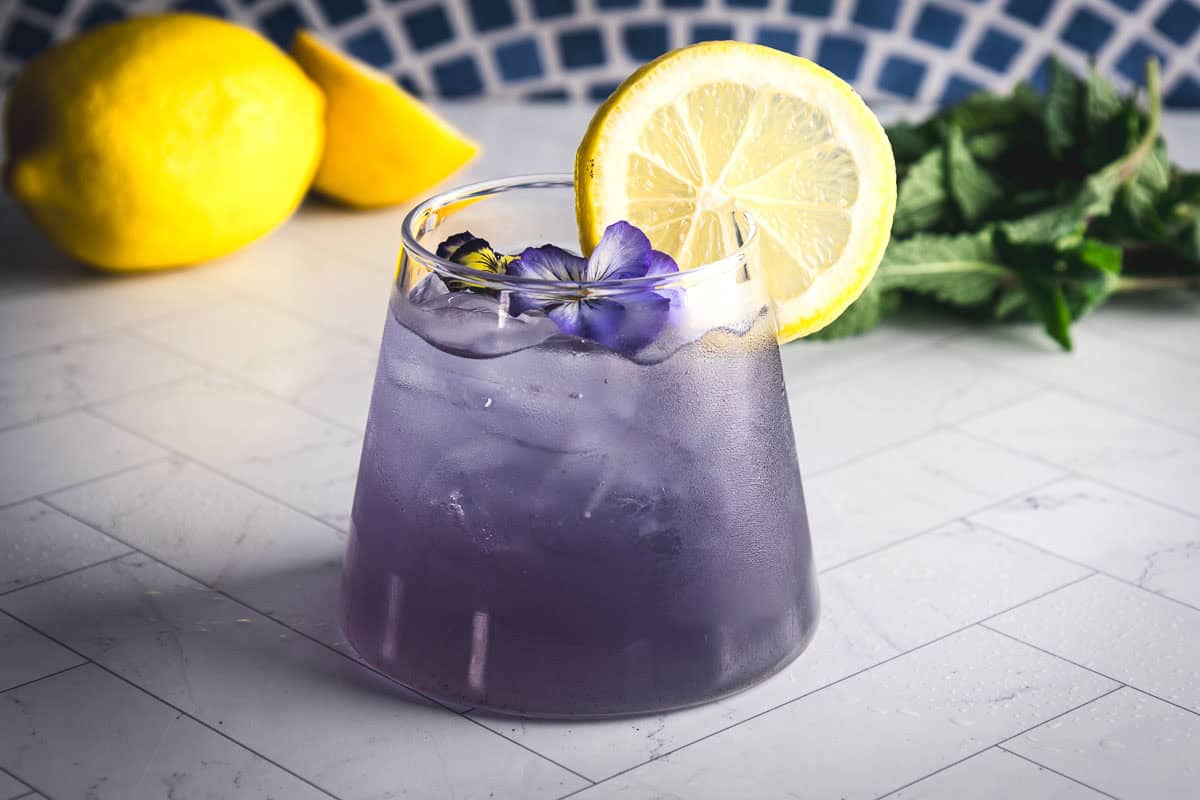 A glass of Purple Electrolyte Mocktail with a lemon slice and purple flower garnish, lemons and mint in the background.