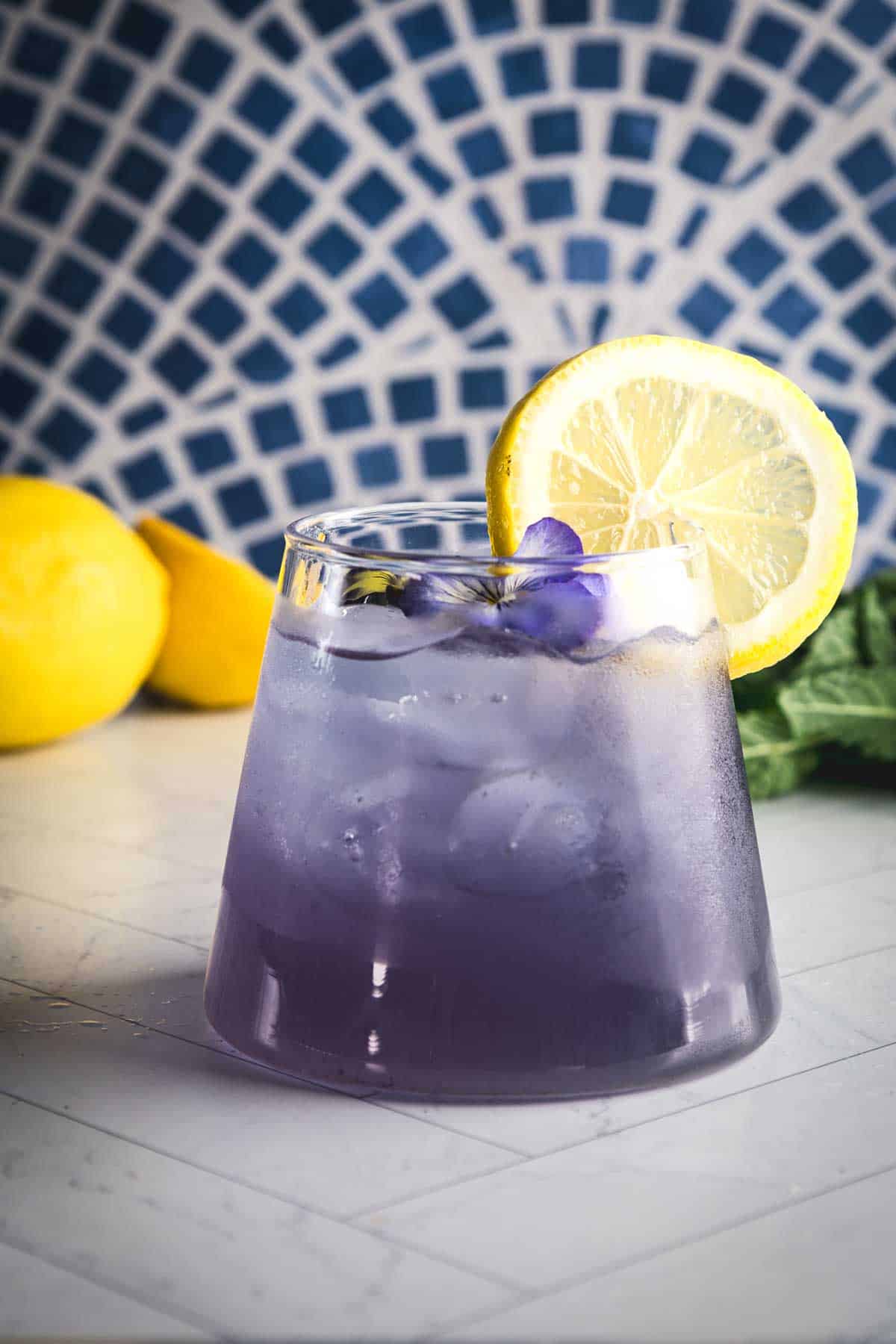 A purple electrolyte mocktail in a glass, topped with a lemon slice and violet petals, with fresh lemons in the background.
