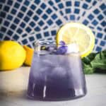 Purple Electrolyte Mocktail with lemon slice garnish, fresh lemons, and a blue mosaic background.