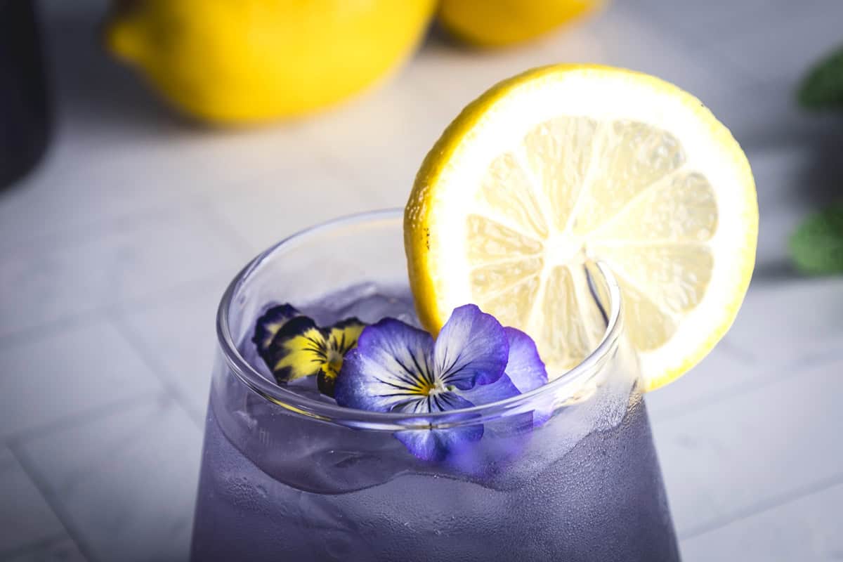 A glass of Purple Electrolyte Mocktail, iced and garnished with a lemon slice and purple edible flowers.