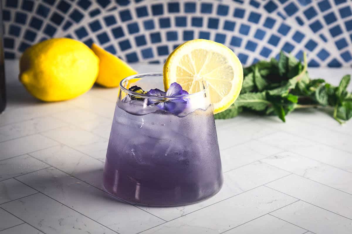 A vibrant purple mocktail garnished with a lemon slice and edible flowers, this refreshing drink is the perfect electrolyte mocktail for any occasion, with fresh lemons and mint in the background.