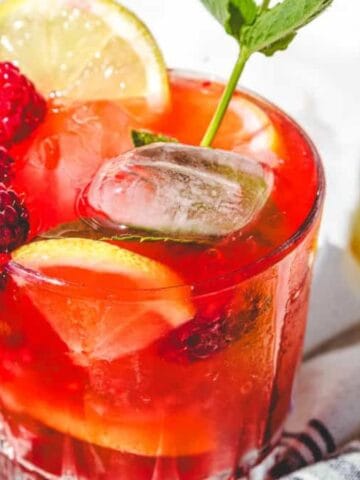 A glass of iced raspberry lemonade garnished with raspberries, lemon slices, and a sprig of mint.