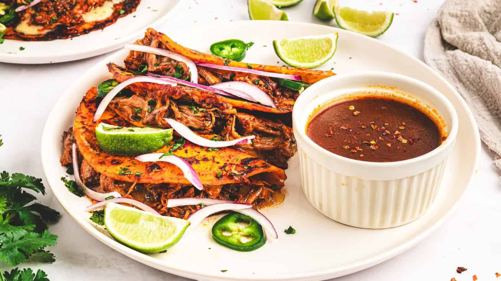 Three birria tacos with lime wedges, sliced onions, and dipping sauce on a white plate.