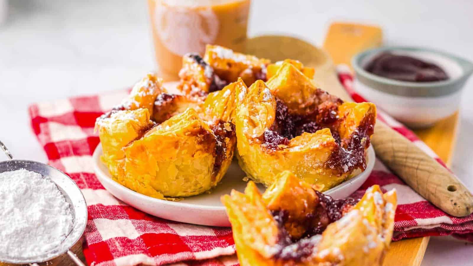 Golden puff pastries with jam filling on a white plate, set on a red checkered cloth with powdered sugar nearby.