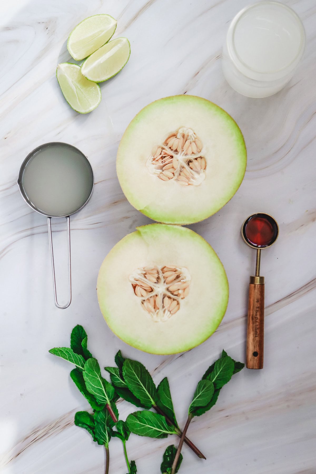 Halved melon, mint leaves, lime wedges, juice in a strainer and glass, and a melon baller on a marble surface&mdash;perfect for preparing a refreshing Melon Margarita Mocktail.