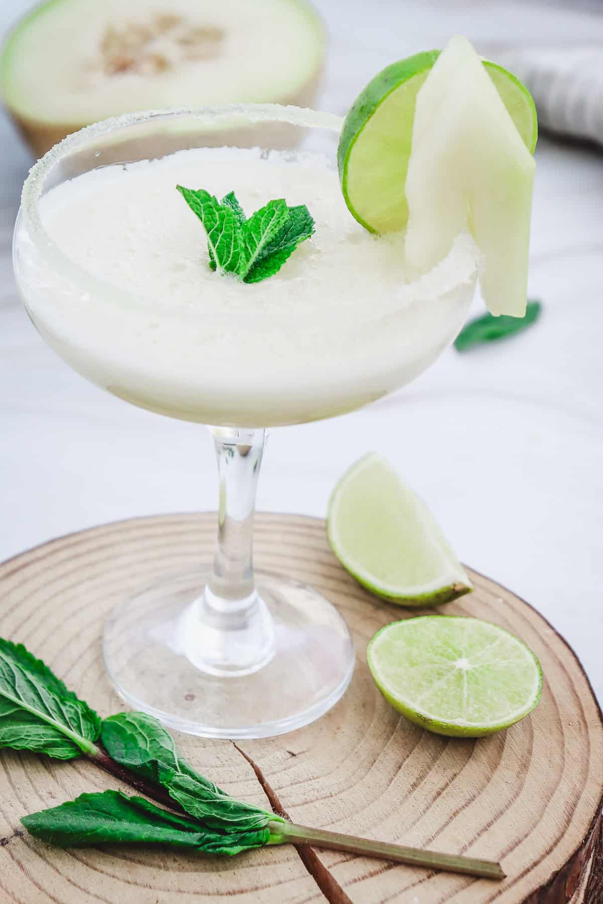 A creamy white Melon Margarita Mocktail in a glass, garnished with lime, melon slice, and fresh mint on a wooden coaster.