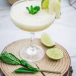 A creamy Melon Margarita Mocktail garnished with mint, lime, and melon slices sits on a wooden coaster.