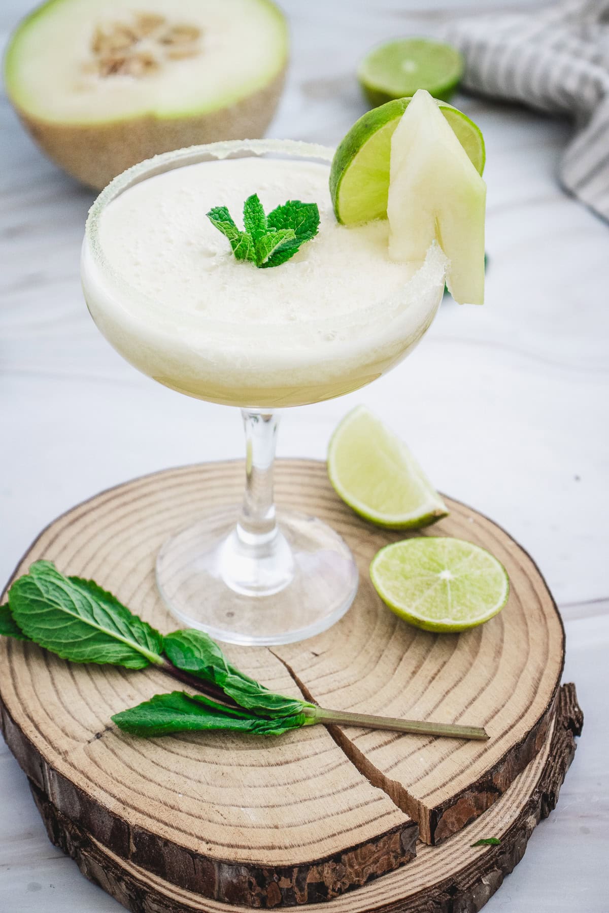 A creamy Melon Margarita Mocktail garnished with mint, lime, and melon slices sits on a wooden coaster.