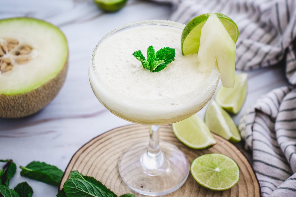A creamy Melon Mocktail in a glass, garnished with mint, lime, and a melon slice, sits on a wooden board&mdash;an inviting mocktail recipe perfect for any occasion.