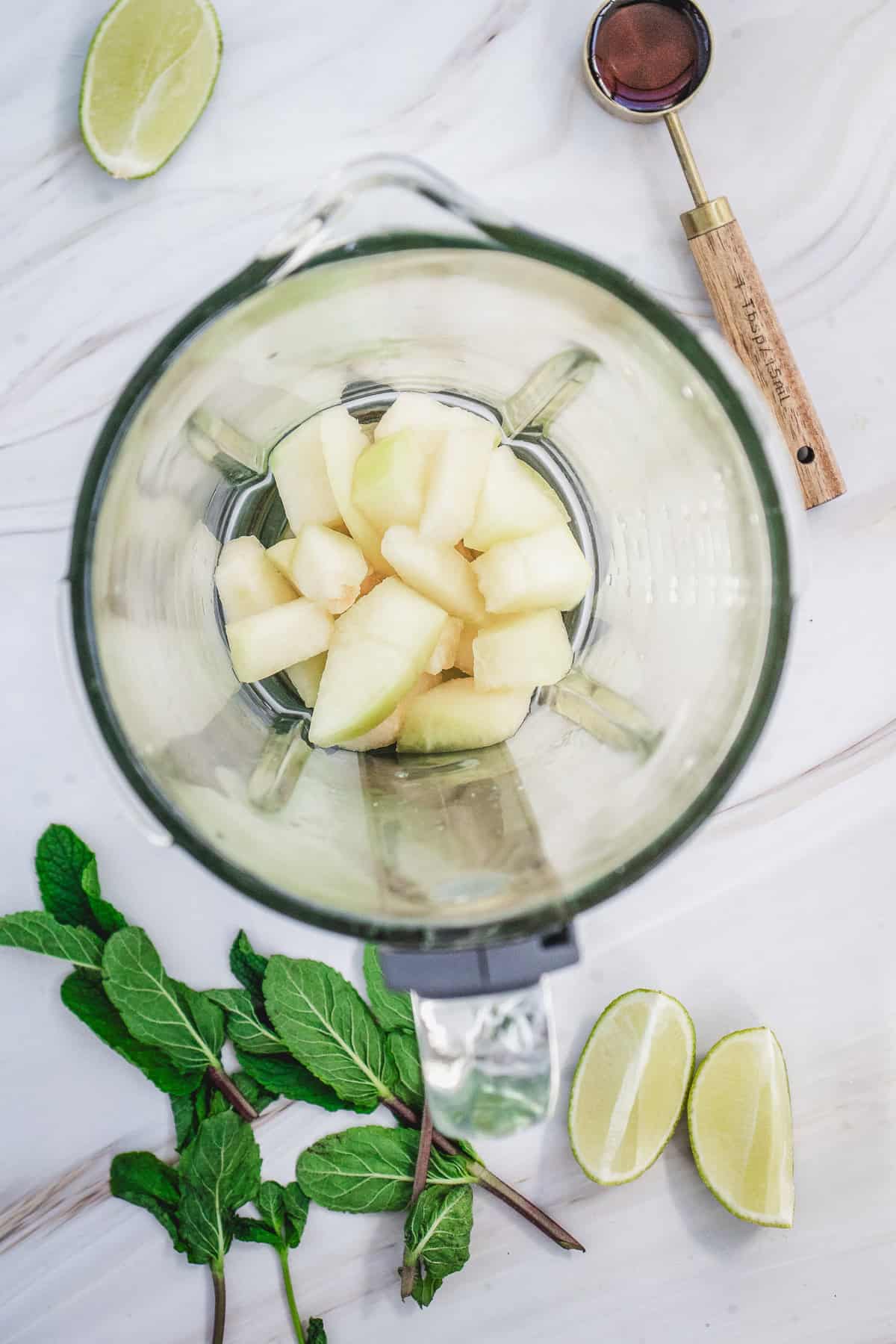 Chunks of melon in a blender, surrounded by lime wedges, mint leaves, and a measuring spoon on a white surface&mdash;perfect ingredients for a refreshing Melon Margarita Mocktail.