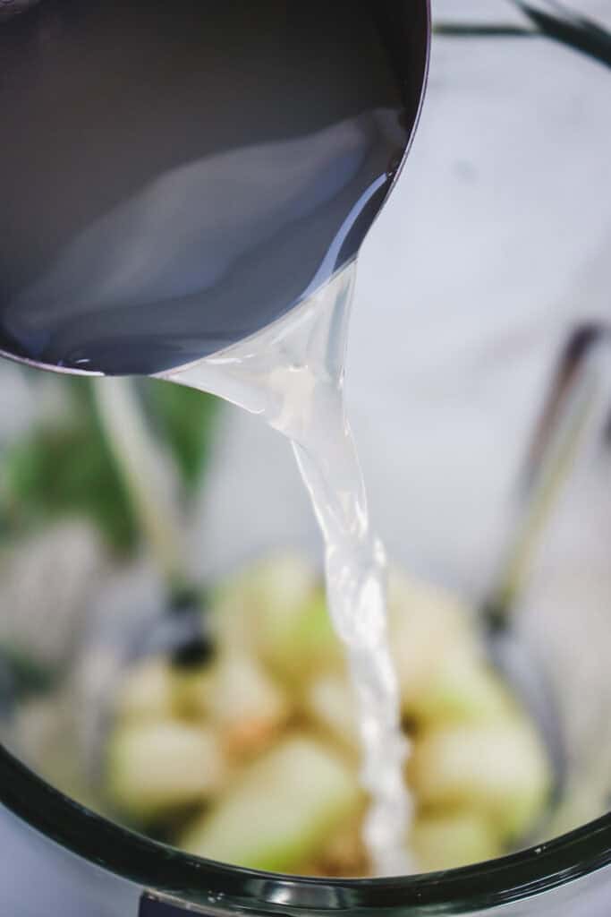 A close-up of liquid being poured from a metal cup into a blender with cut fruit inside, perfect for crafting a refreshing Melon Margarita Mocktail.