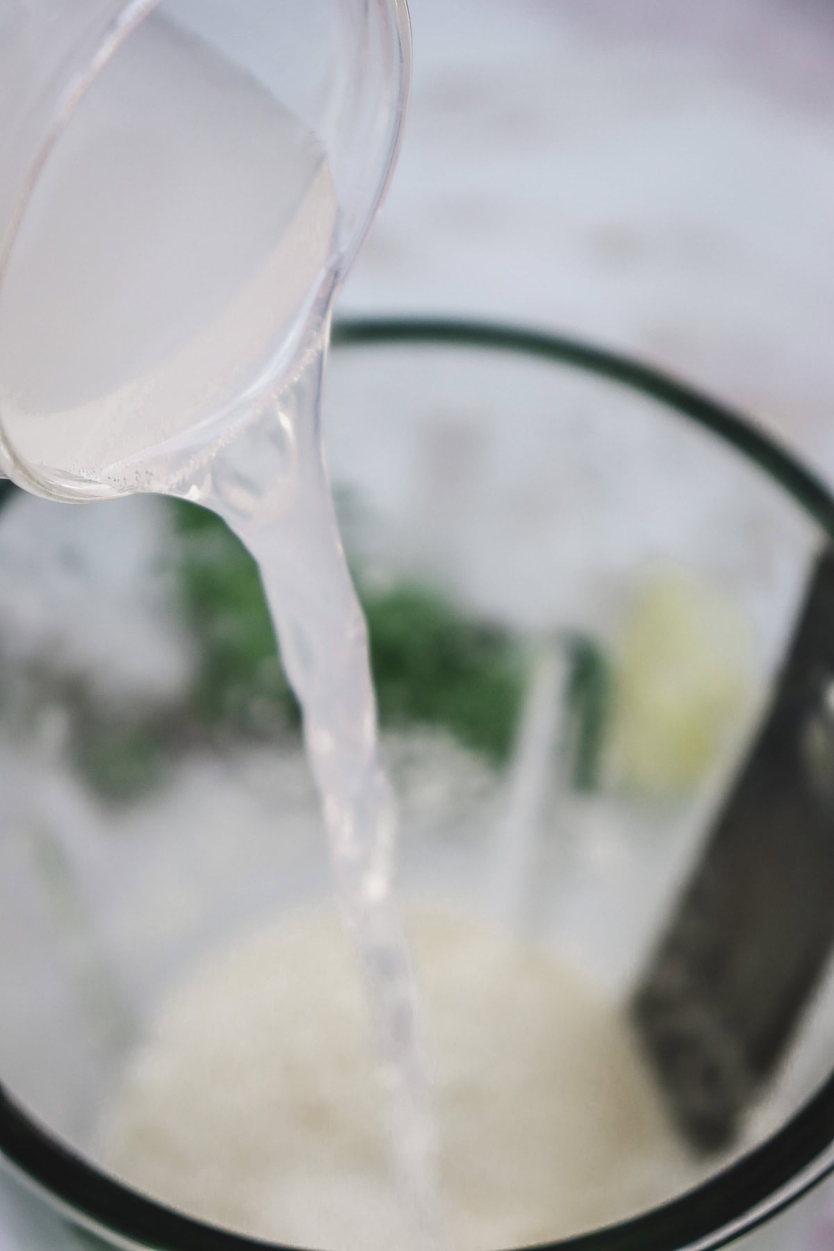 A clear liquid, perfect for a melon margarita mocktail recipe, is being poured from a glass into a mixing bowl with a whisk inside.