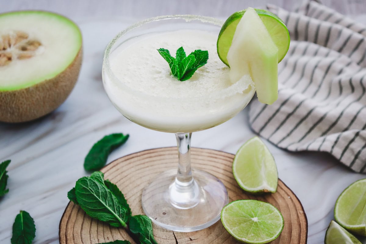 A melon margarita mocktail in a glass, garnished with lime, melon slice, and mint, surrounded by fresh limes and mint leaves.