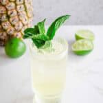 A Tropical Electrolyte Mocktail garnished with mint sits next to a pineapple and sliced limes on a white surface.