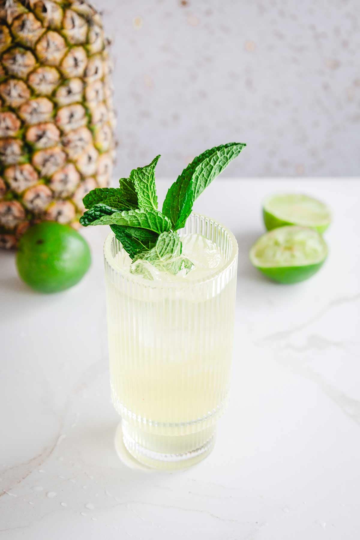 A Tropical Electrolyte Mocktail garnished with mint sits next to a pineapple and sliced limes on a white surface.