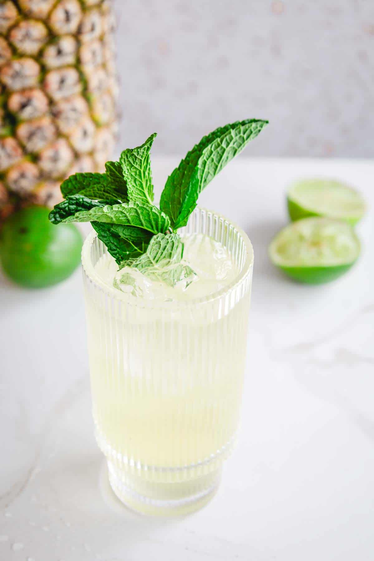 A glass of light yellow Tropical Electrolyte Mocktail with ice, garnished with fresh mint, sits beside limes and a pineapple.