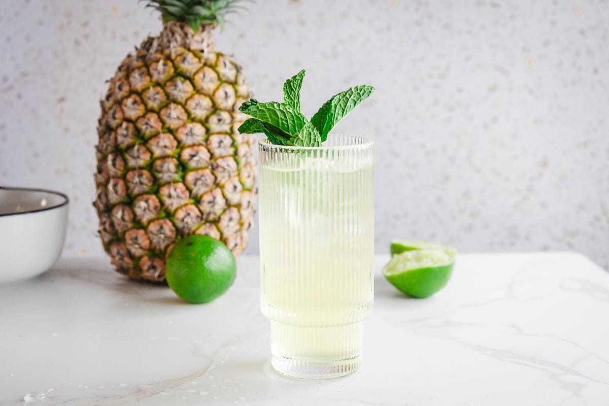 A glass of yellow-green tropical mocktail with mint, next to a whole pineapple and limes on a white surface.