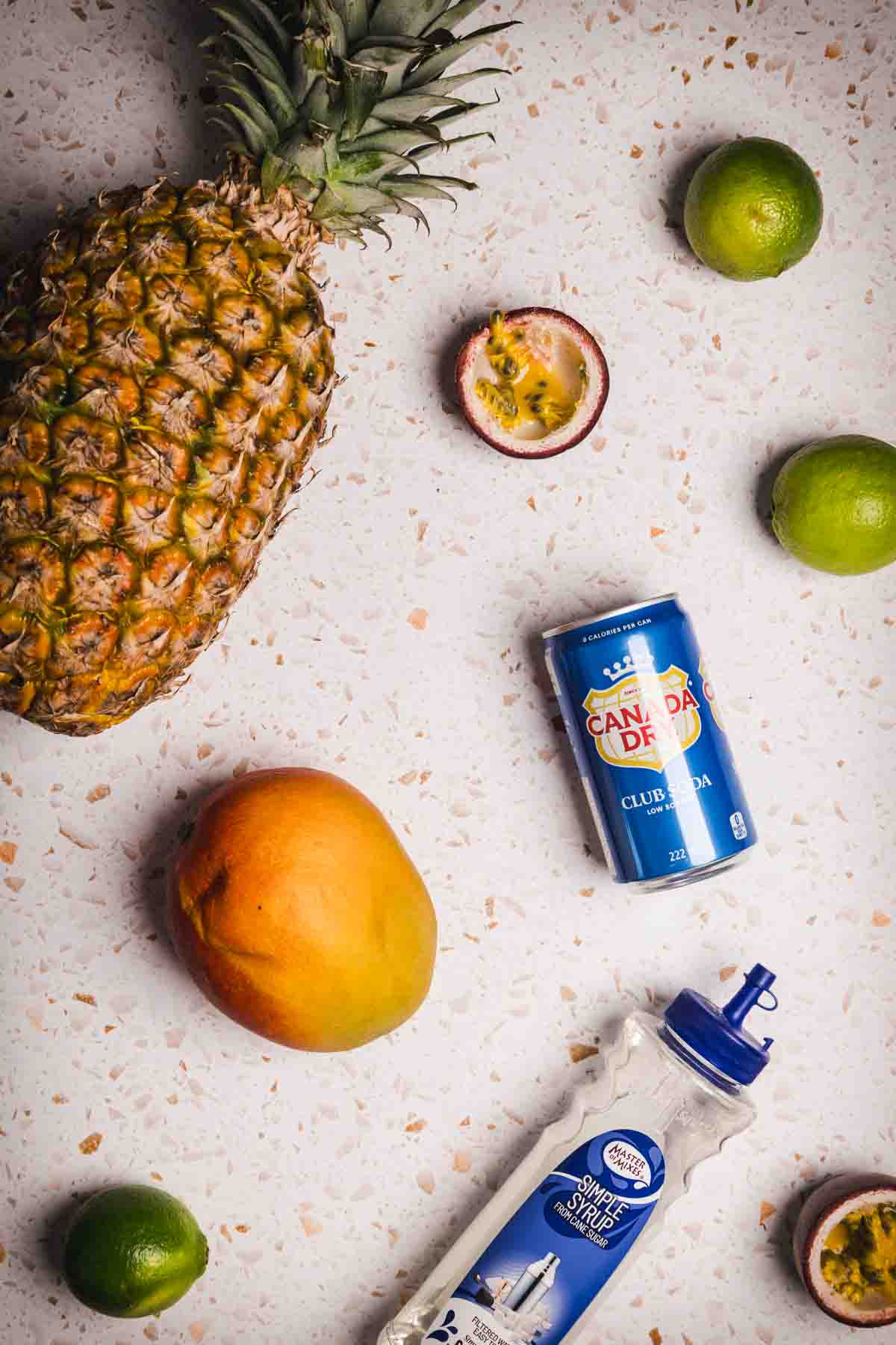 Pineapple, mango, limes, passionfruit, a can of ginger ale, and a club soda bottle on a white surface&mdash;perfect ingredients for a refreshing Tropical Mocktail.
