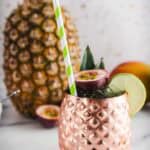 A vibrant tropical mocktail served in a copper pineapple cup with a striped straw, lime slice, and passionfruit garnish.