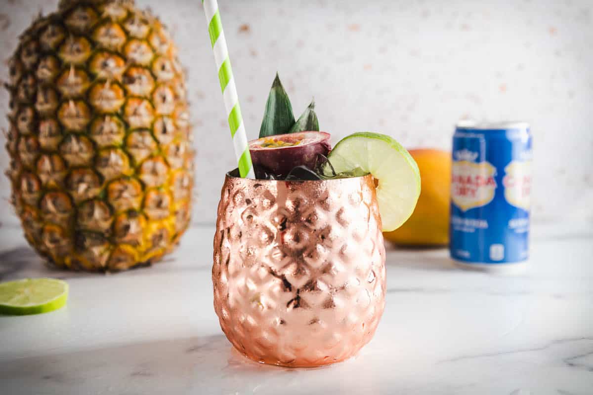 Copper pineapple cup with a striped straw, lime slice, and fruit garnish&mdash;perfect for serving your favorite Tropical Mocktail; pineapple in background.