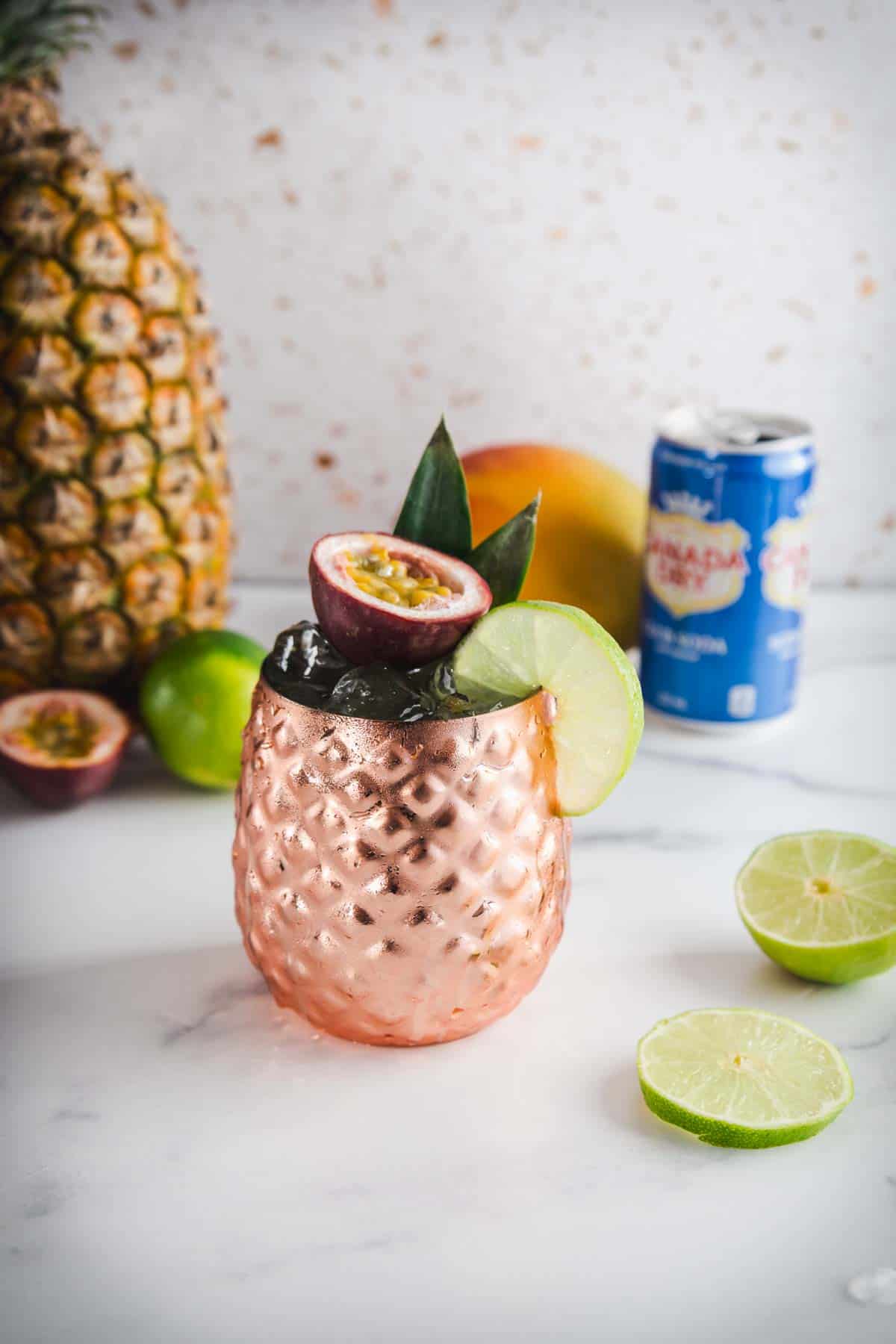 A copper mug with a lime slice and passionfruit, surrounded by tropical fruits and a soda can&mdash;perfect for serving your favorite Tropical Mocktail.