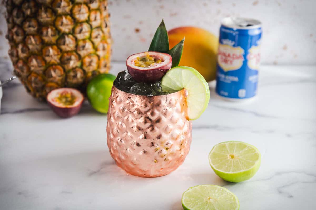 A copper pineapple cup with a tropical drink, garnished with passionfruit, lime, and pineapple leaves&mdash;perfect for showcasing your favorite Tropical Mocktail Recipe.
