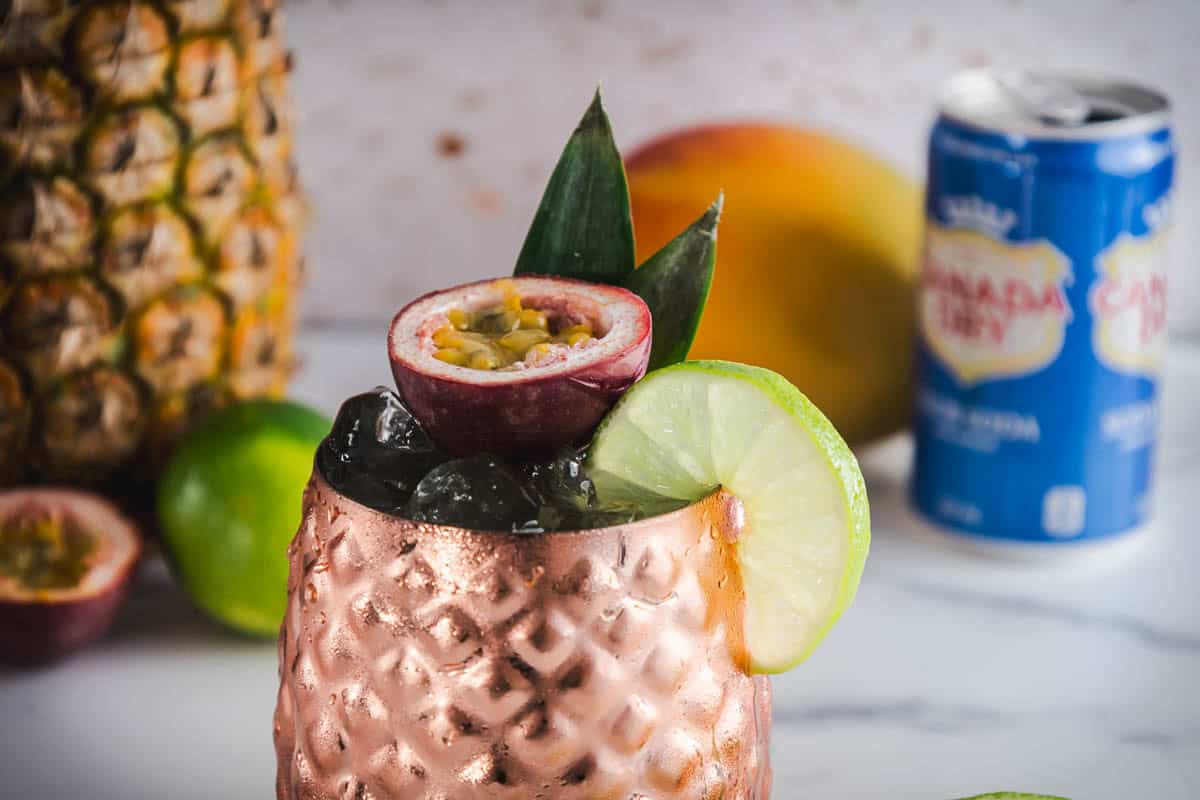 A copper cup with ice, garnished with lime and passionfruit, sits near pineapple, mango, and a soda can&mdash;perfect for a refreshing Tropical Mocktail.