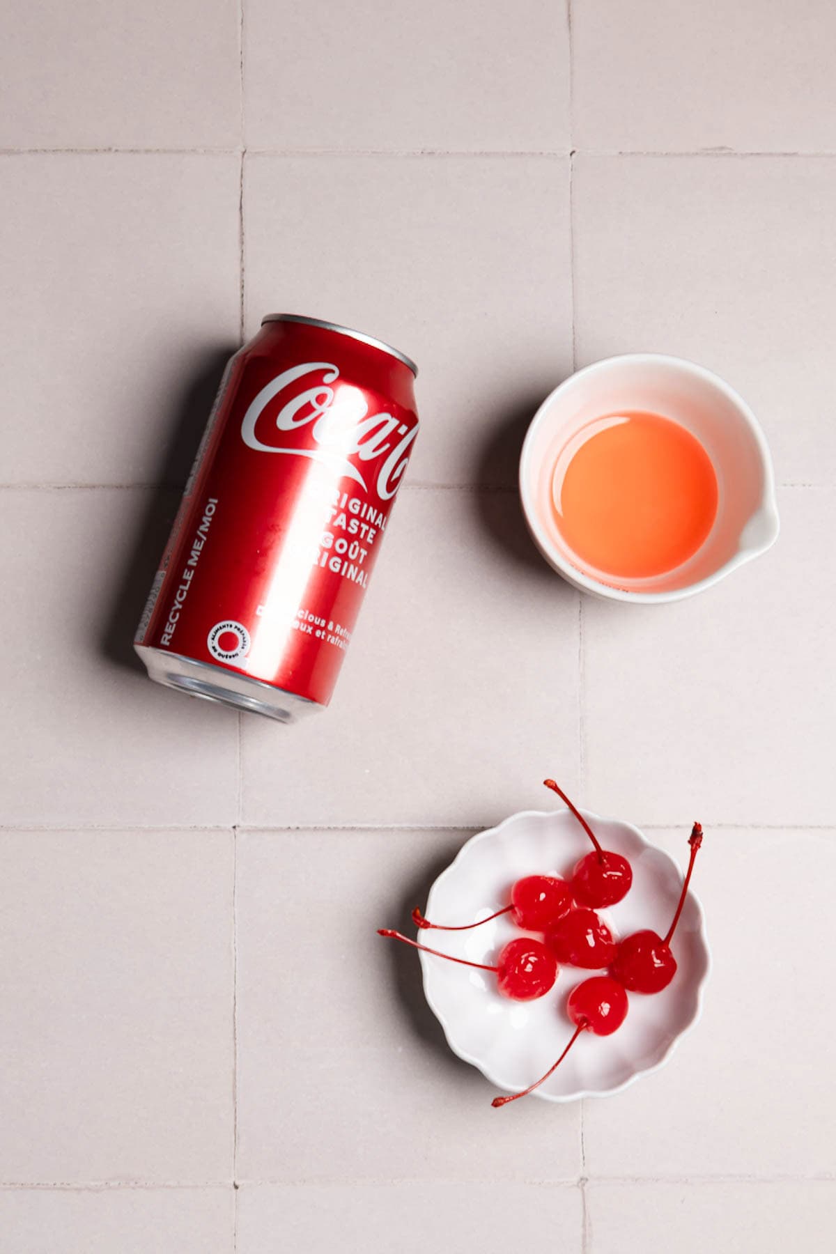 A can of Coca-Cola, a small cup of pink liquid, and a bowl of maraschino cherries on a tiled surface&mdash;perfect ingredients for crafting a classic Roy Rogers cocktail.