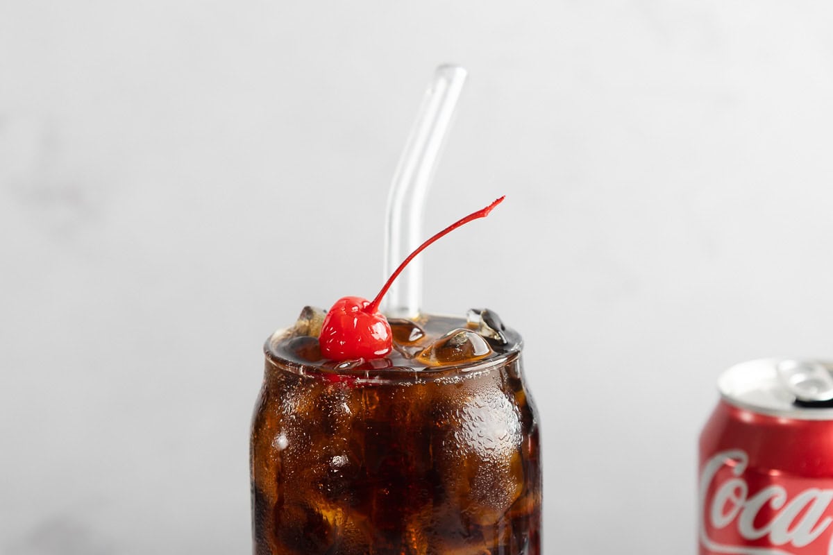 A glass of cola with ice, a cherry on top, a clear straw, and a Coca-Cola can beside it&mdash;just like the classic Roy Rogers cocktail and vintage drink recipe.