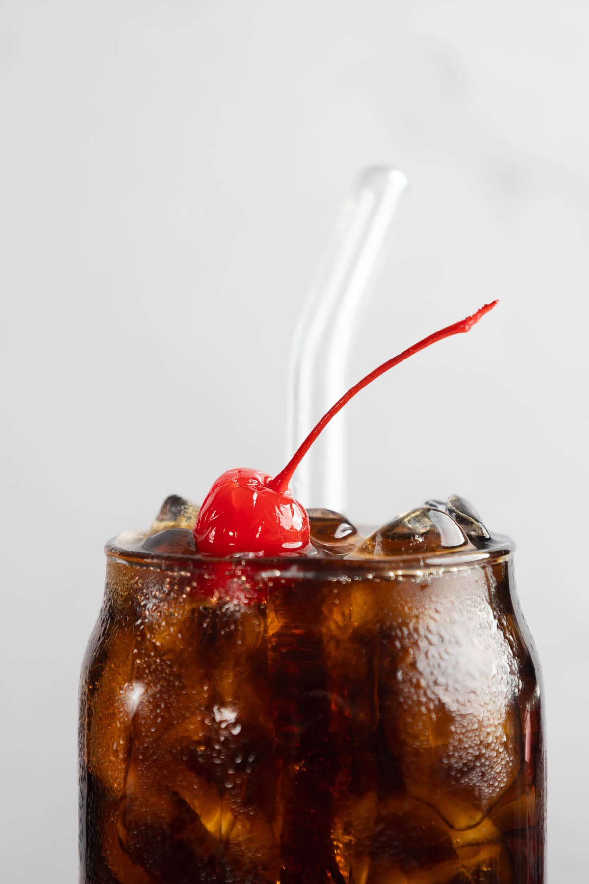 A glass of iced cola with a red cherry on top and a clear straw evokes the classic Roy Rogers, a vintage drink recipe that&rsquo;s the perfect non-alcoholic cocktail for any occasion, set against a light background.