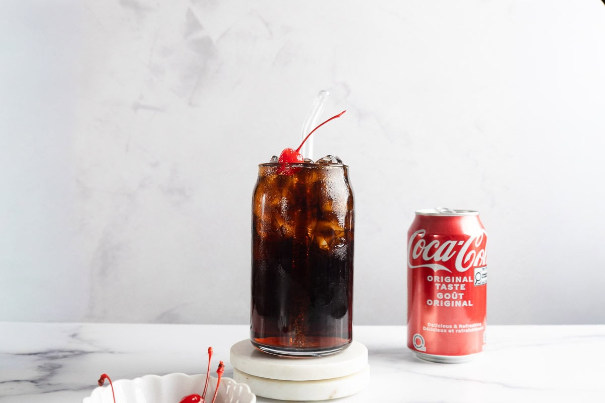 A glass of iced cola with a cherry on top, inspired by the classic Roy Rogers vintage drink, sits beside a can of Coca-Cola on a white surface.