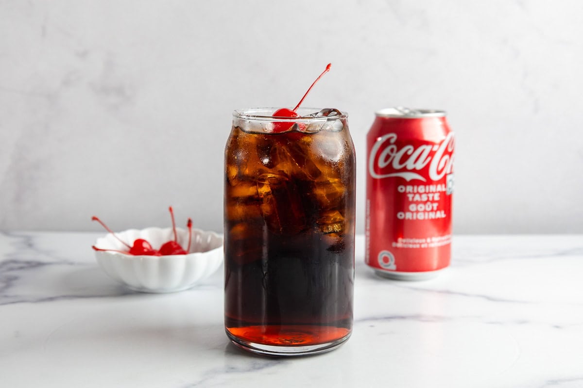 A glass of iced cola with cherries, a can of Coca-Cola, and a bowl of cherries on a white surface evoke vintage charm&mdash;perfect for creating a classic Roy Rogers drink recipe.