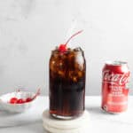 A vintage glass of cola with ice and a cherry, next to a can of Coca-Cola and a bowl of cherries—just like the classic Roy Rogers drink recipe.