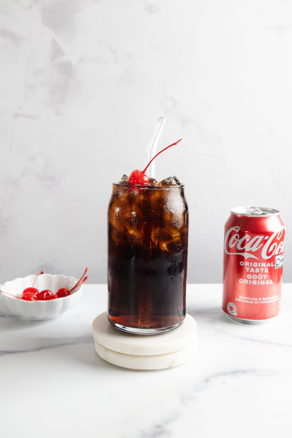 A vintage glass of cola with ice and a cherry, next to a can of Coca-Cola and a bowl of cherries&mdash;just like the classic Roy Rogers drink recipe.