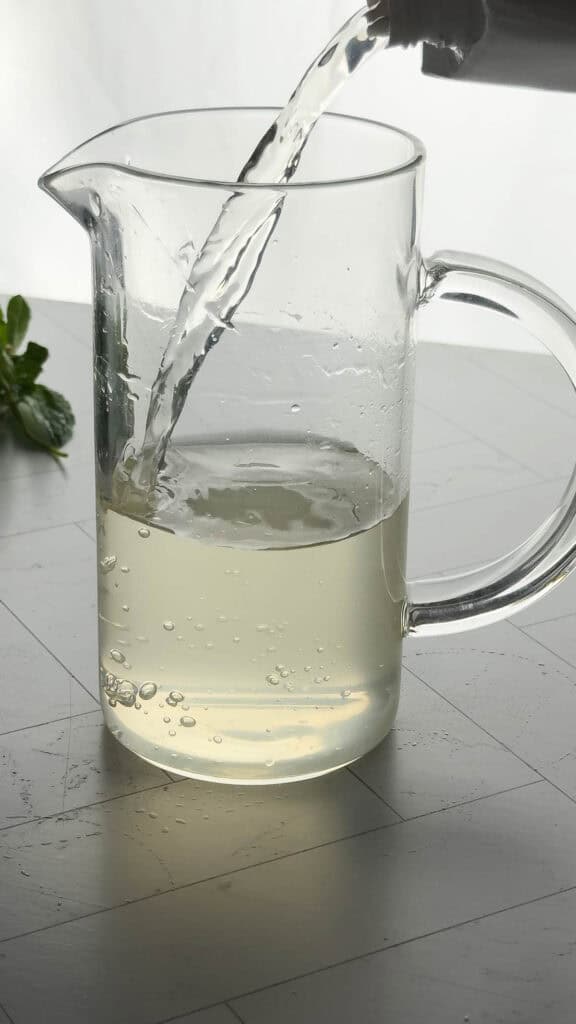 A glass pitcher being filled with a light yellow liquid for a refreshing electrolyte mocktail, with mint leaves in the background.